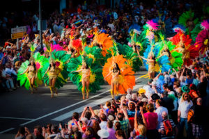 Živý pouličný sprievod počas karnevalu s davmi ľudí sledujúcich mužov a ženy v pestrofarebných kostýmoch tancujúcich na rytmy bubnov.