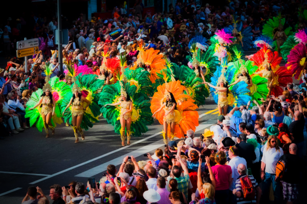 Défilé de rue animé lors du carnaval, avec des foules qui regardent des hommes et des femmes en costumes colorés danser au rythme des tambours.